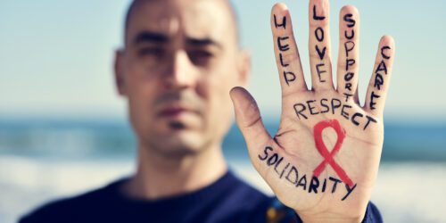 closeup of the hand of a young caucasian man with a red ribbon for the fight against AIDS painted in it, and the words helps, love, support, care, respect and solidarity