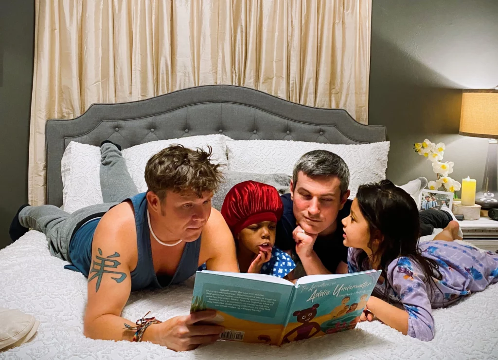 Two dads sitting on their bed reading a book with their two daughters