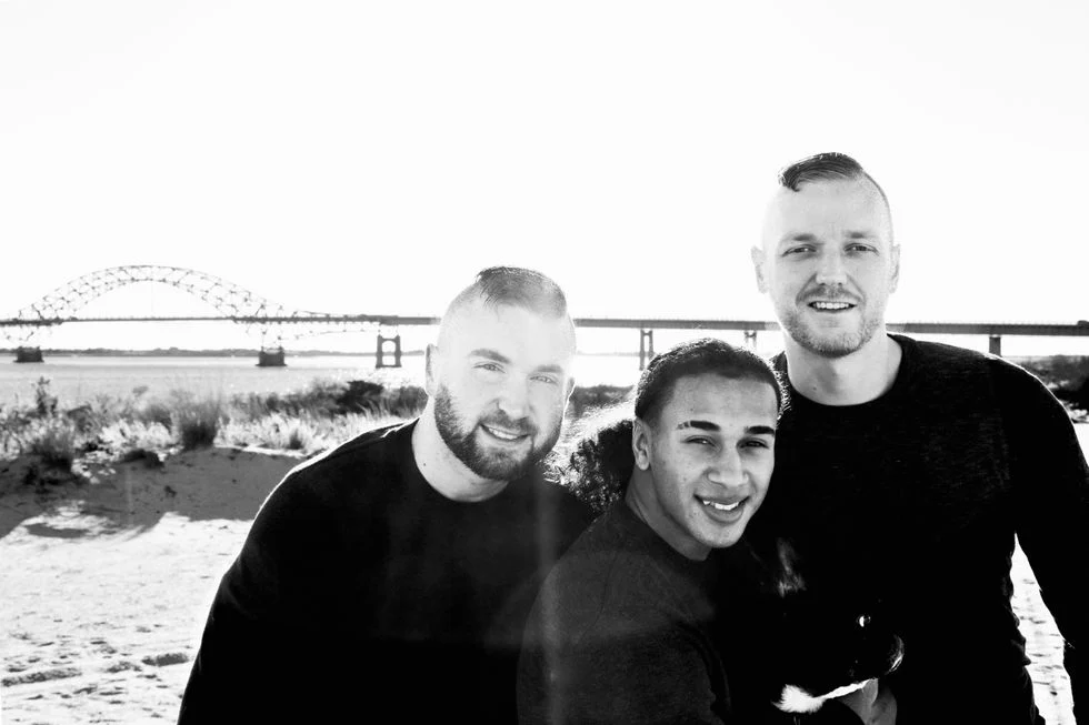 Black and white photo of two foster dads posing with their son in front of a bridge