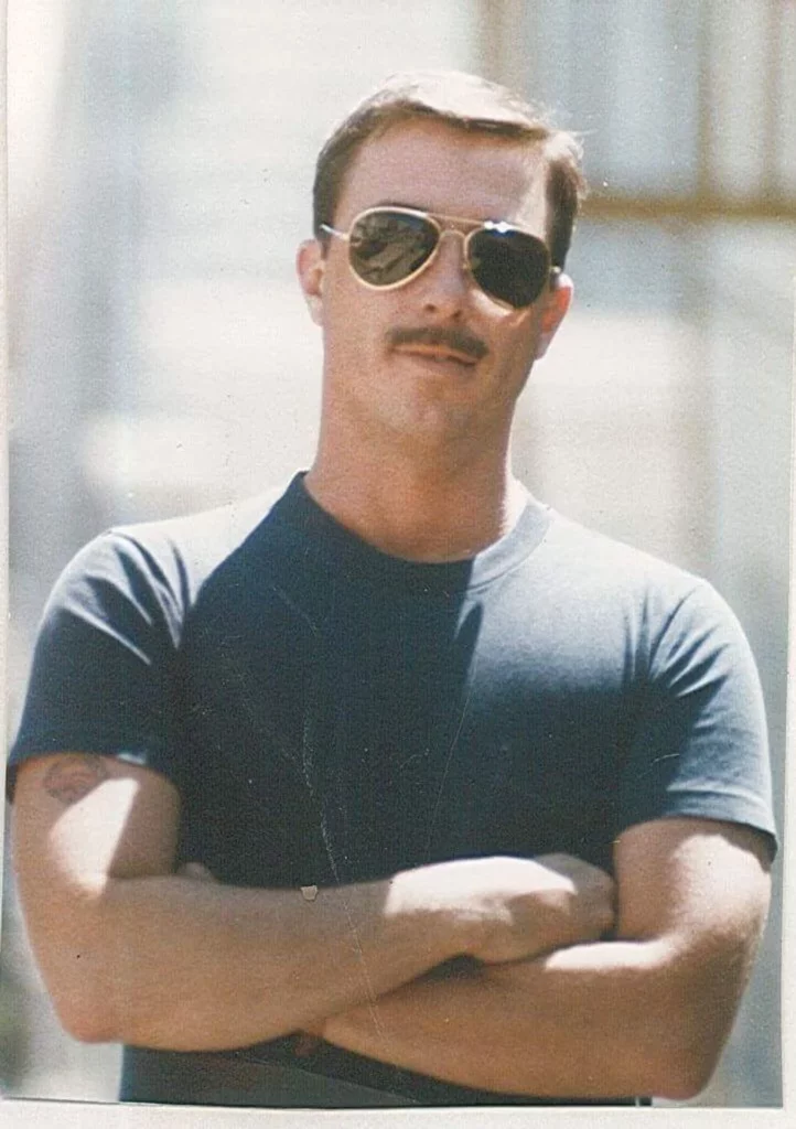 Man with a moustache standing with arms folded wearing aviator sunglasses