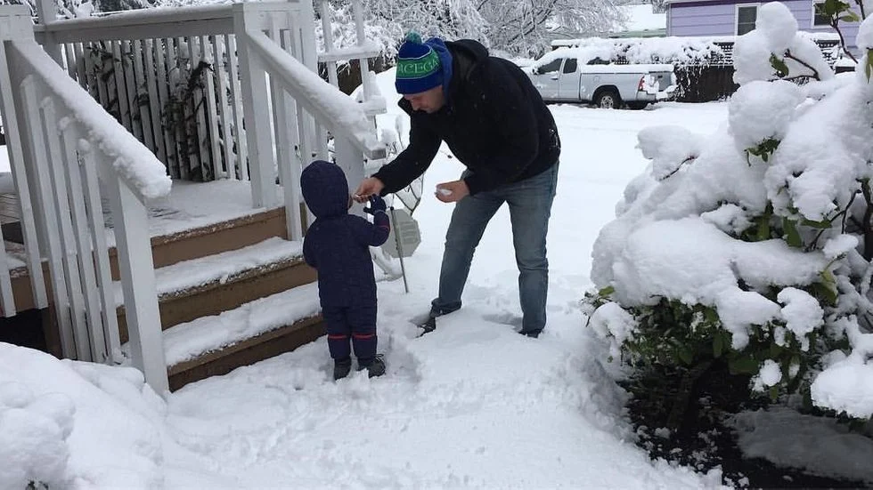 Man with a toddler in a snow suit out in the front yard playing in snow