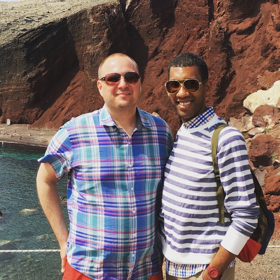 Interracial gay couple posing on a beach with a cliff behind them.