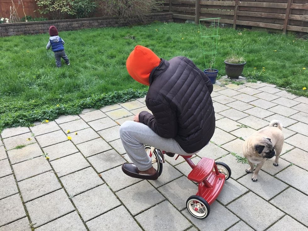 Man riding a little red tricycle in a backyard with a toddler