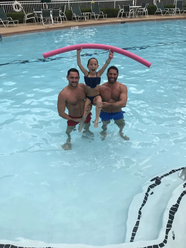 Two dads in the pool with their daughter holding a pool noodle