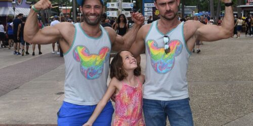 Two dads in Mickey Mouse shirts flexing their muscles with their daughter in betwen them
