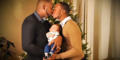 Gay couple kissing in front of their Christmas tree with their newborn