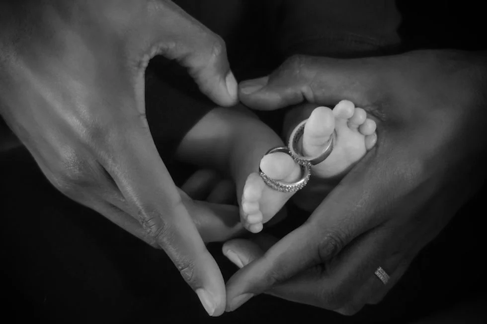 Two mens hands forming heart around baby's feet with wedding rings