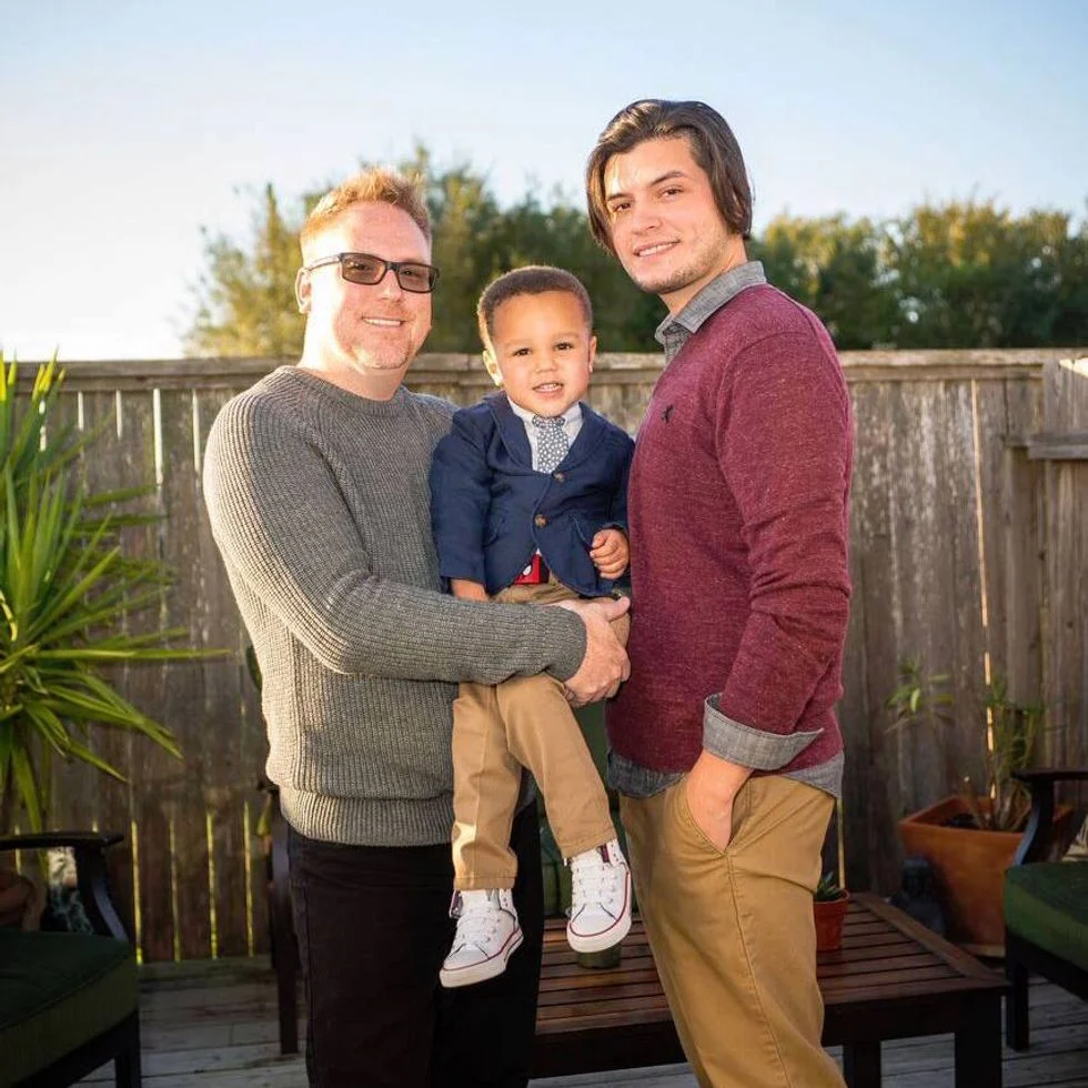 Two foster dads holding their son outside.