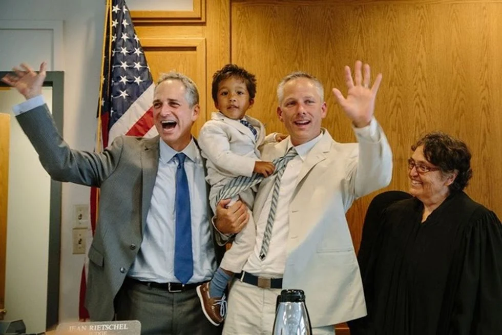 Two foster dads holding their son and celebrating adoption day with the judge in the courtroom