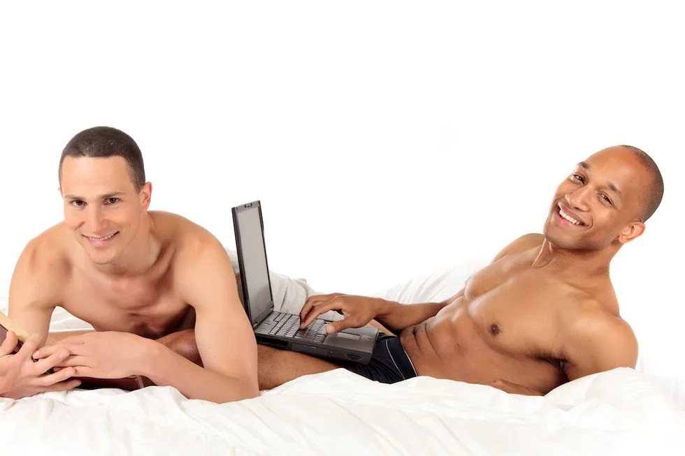 Gay couple lying on their bed shirtless. One with a laptop