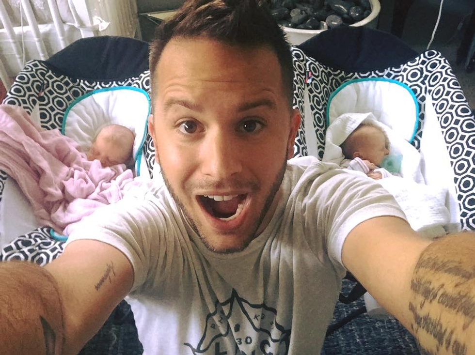 Dad taking a selfie when he is standing in-between two cribs with his newborn twins in them