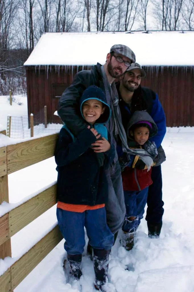 Two dads standing outside of their barn in the snow with their sons