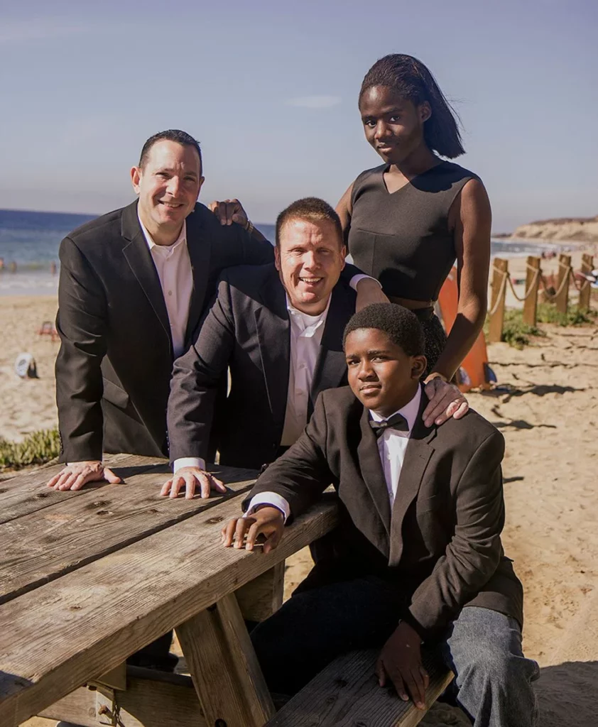 Two dads with their two children dressed in black tie at a picnic table on the beach