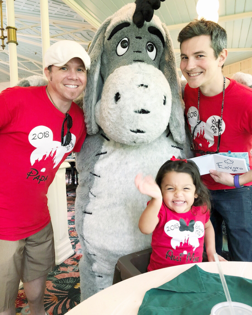 Two dads posing at Disney with their daughter and Eyore
