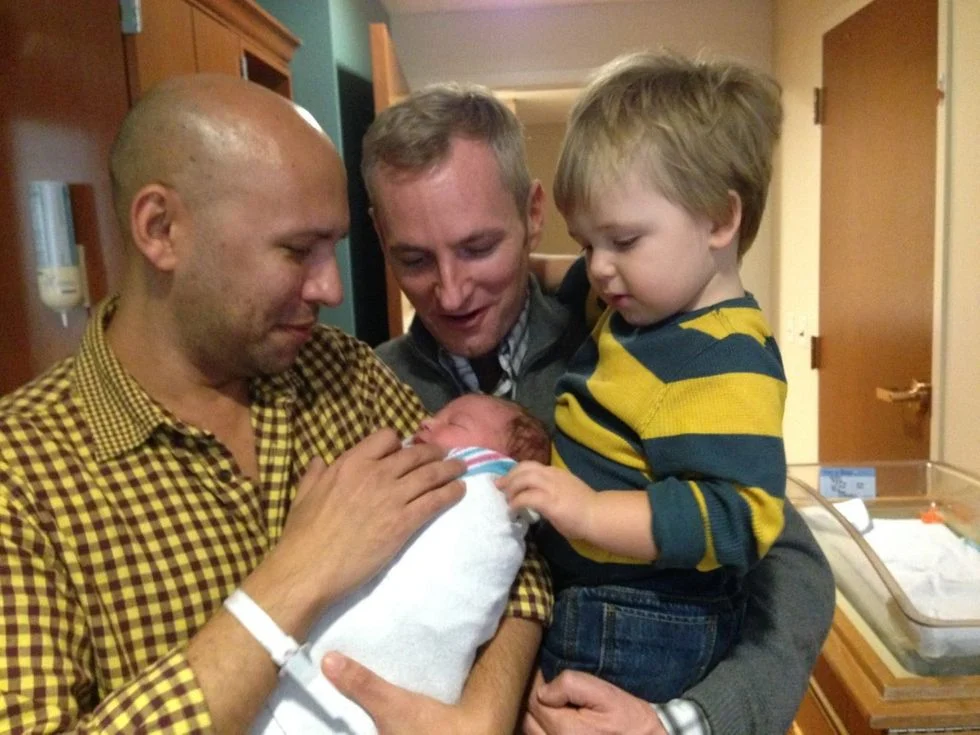 Two dad in the hospital one is holidng their newborn son the other holding their toddler