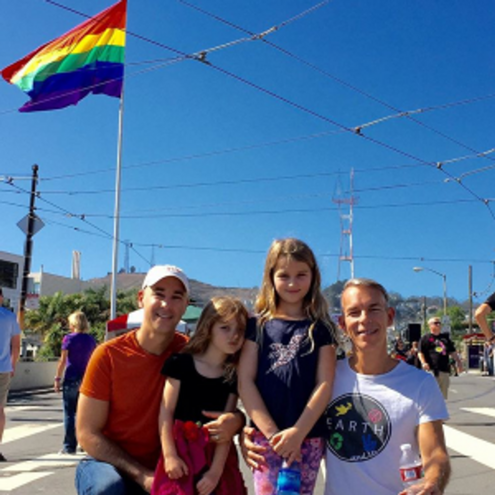 two fathers with their two daughters posing outside under the pride flag