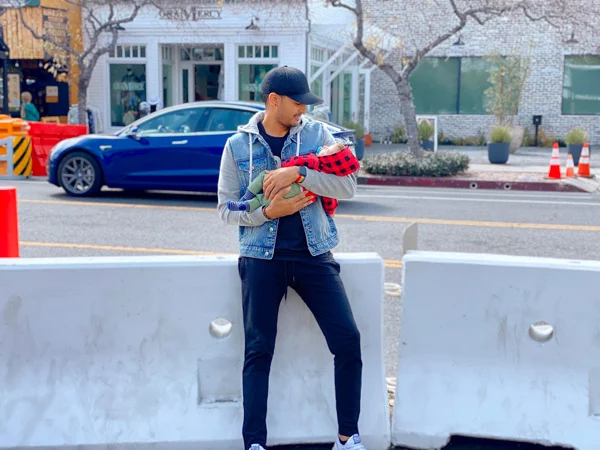 Man wearing a hat holding his newborn son while standing on the sidewalk