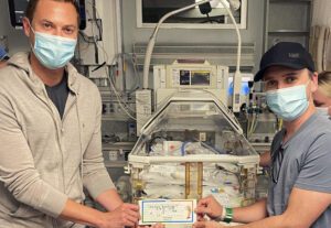 Gay dads hold name sign in front of their premature daughter's crib