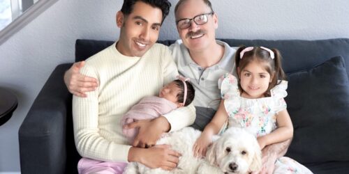 Two dads on the counch with their infant baby, and young daughter, along with their dog